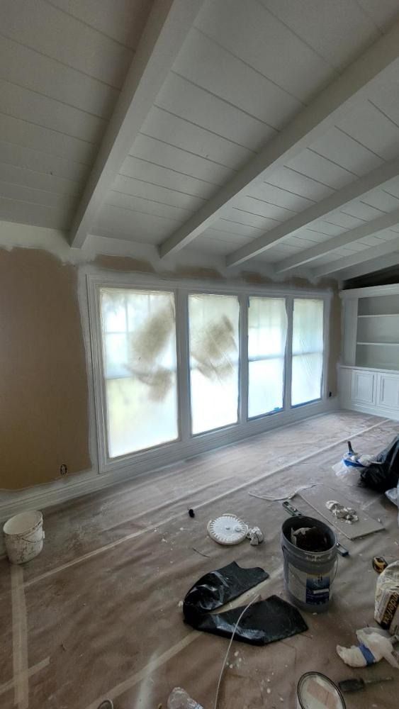 Room being painted: white ceiling with beams, windows covered, tan wall, paint supplies on floor.