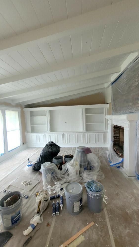 Interior renovation: White-painted ceiling with beams, built-in shelves, fireplace. Painting supplies on floor.