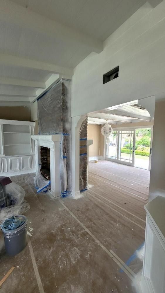 Interior room under renovation, white walls, fireplace covered, wooden floor, archway leads to bright exterior.