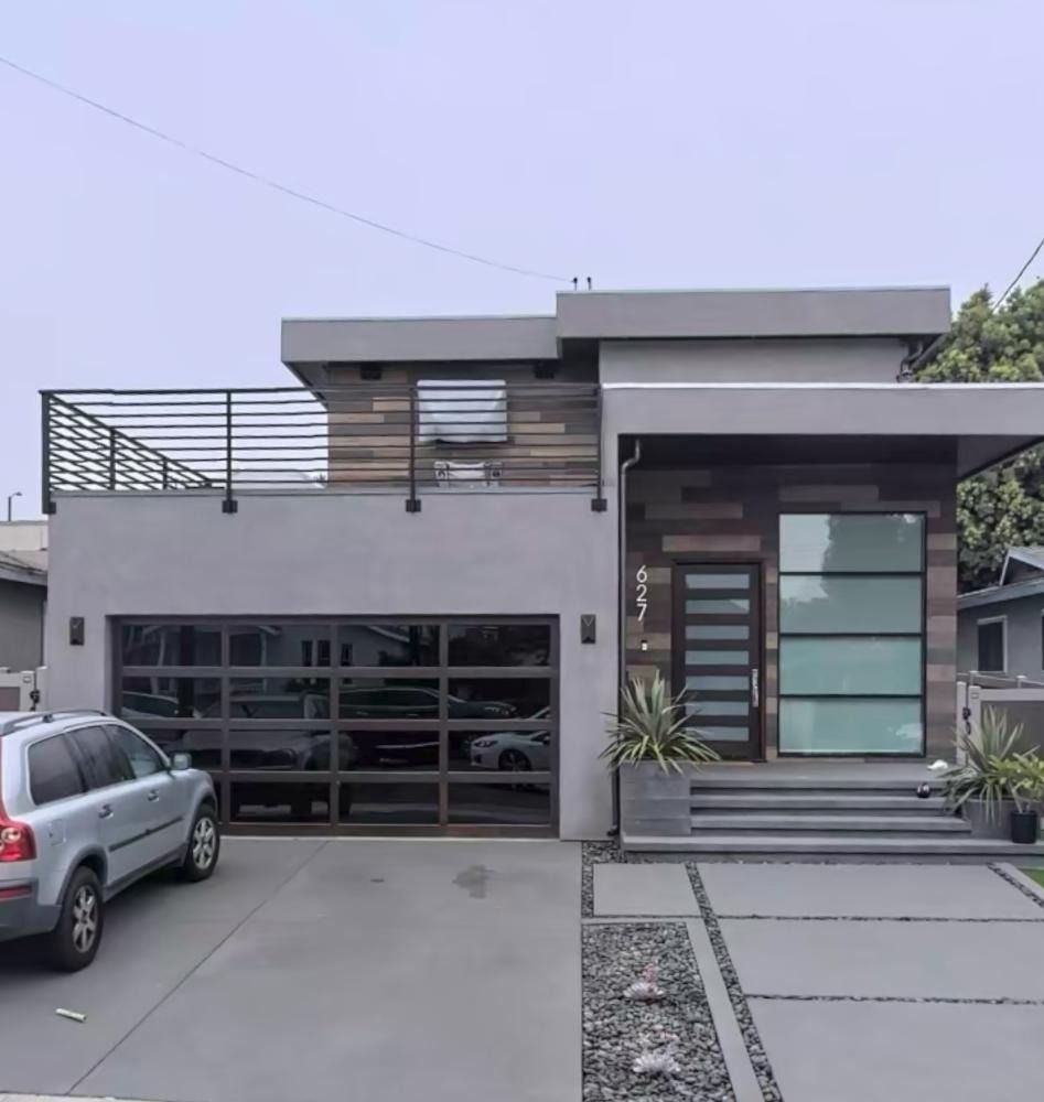 Modern gray house with glass garage door, wood accent, and black railing. SUV parked in driveway.