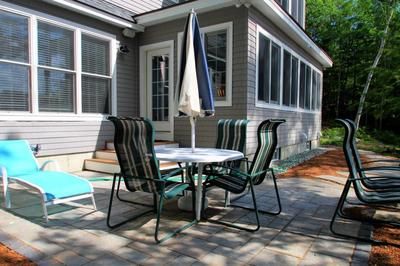 Patio with table, chairs, and umbrella next to a house. Blue chair, grey siding. Sunny day.