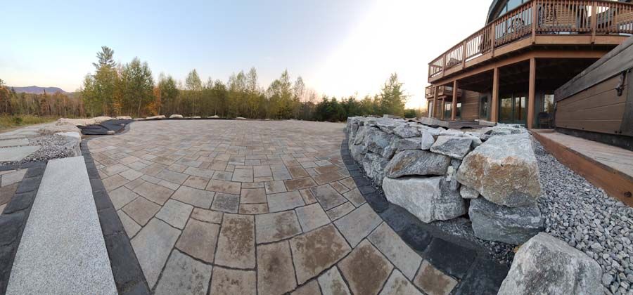 A stone patio with a low retaining wall, leading to a deck, with trees in the background.