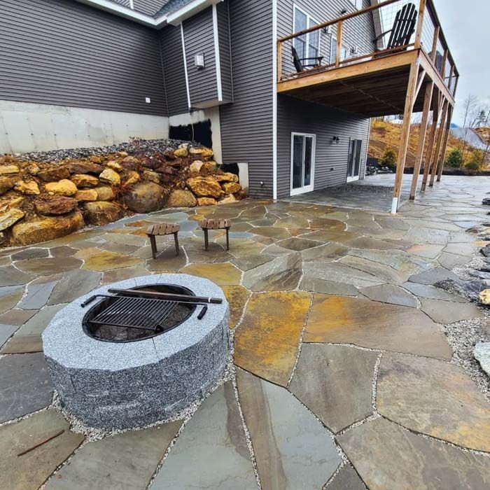 Stone patio with fire pit, two stools, and a deck next to a house.