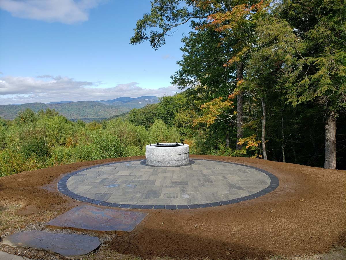 Circular stone patio with fire pit, overlooking mountains and trees under a blue sky.