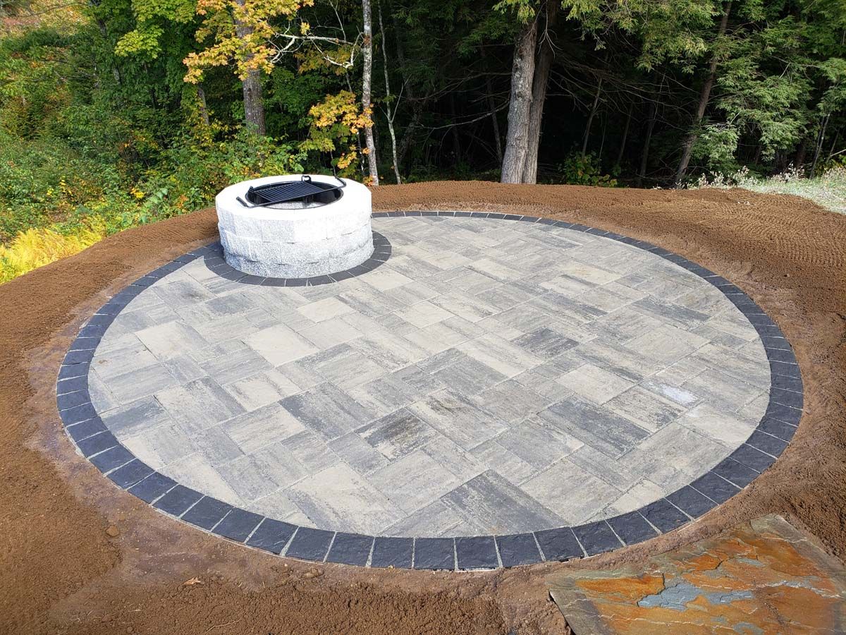 Circular stone patio with fire pit, bordered by dark pavers and brown soil, set in a yard.