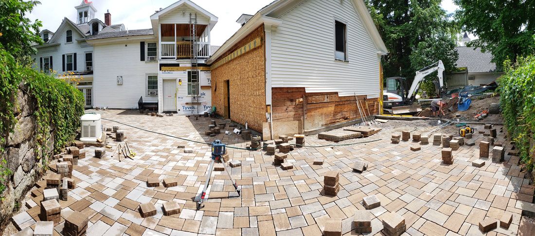 Construction site: laying brick pavers around a building; workers, machinery, and partially built structures are visible.