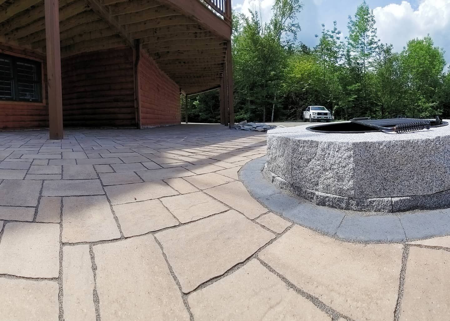 Patio with stone pavers, fire pit, and a car under a wooden deck, surrounded by trees.