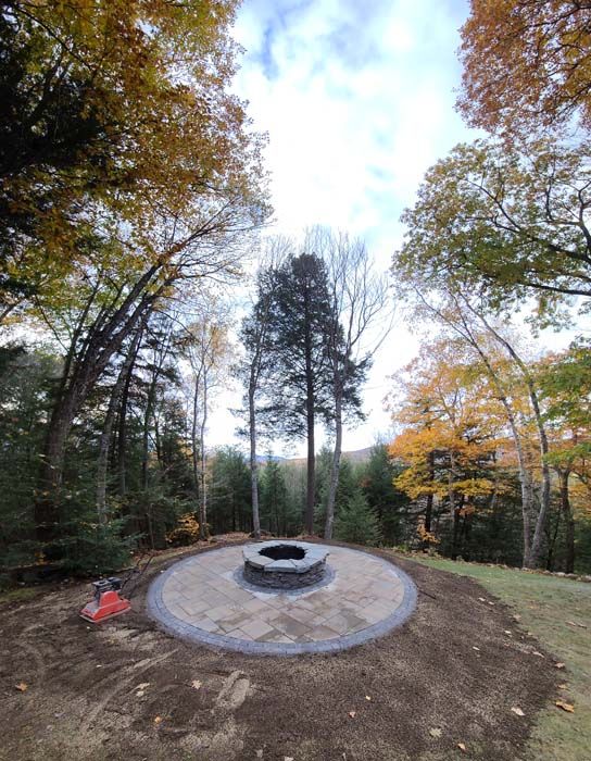 Fire pit surrounded by a circular stone patio, set in a forest with autumn foliage.