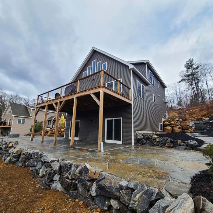 Two-story house with a deck, stone patio, and retaining wall, set in a wooded area under a cloudy sky.
