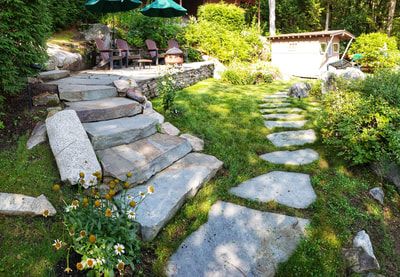 Stone steps and path leading to a lakeside patio with chairs and a small building, surrounded by greenery.