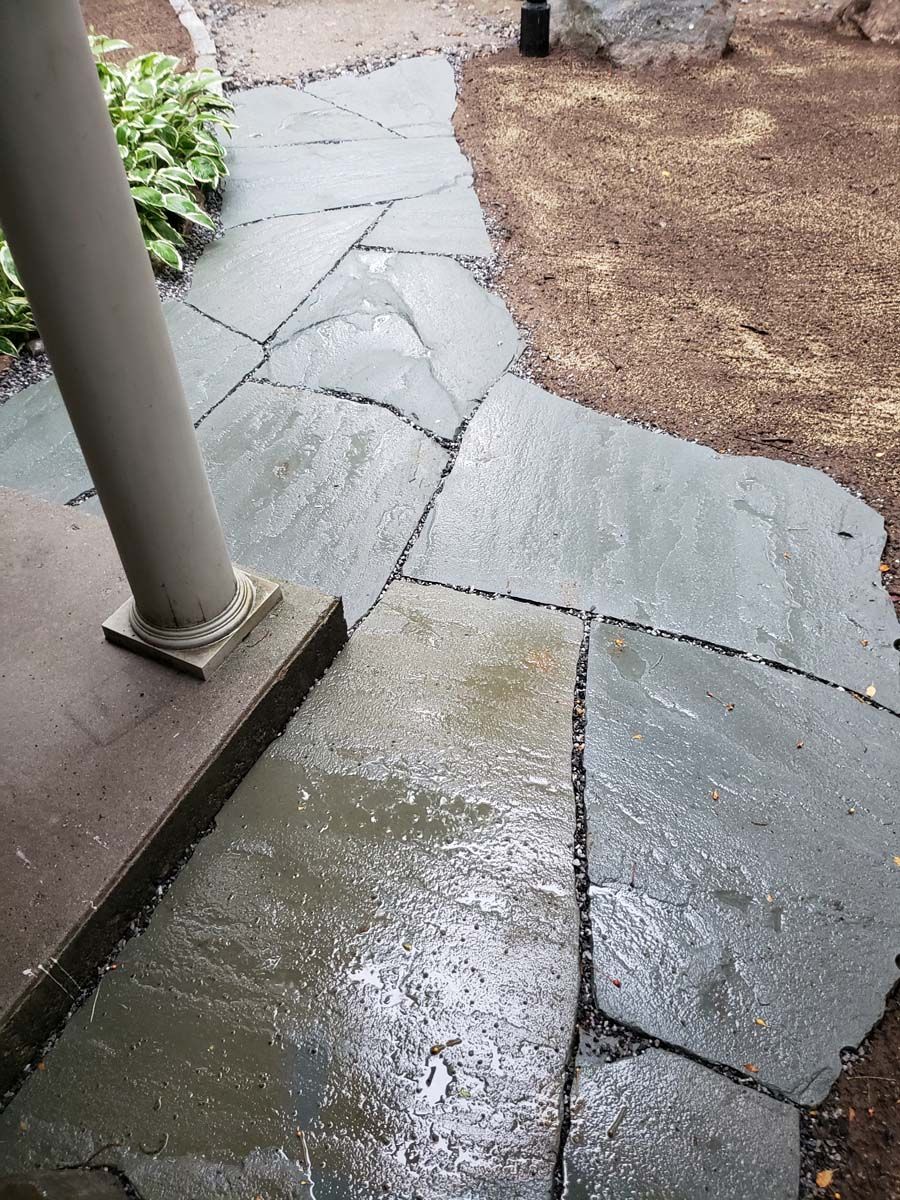 Stone pathway, wet from rain, leads from a covered area, next to a garden and dirt.