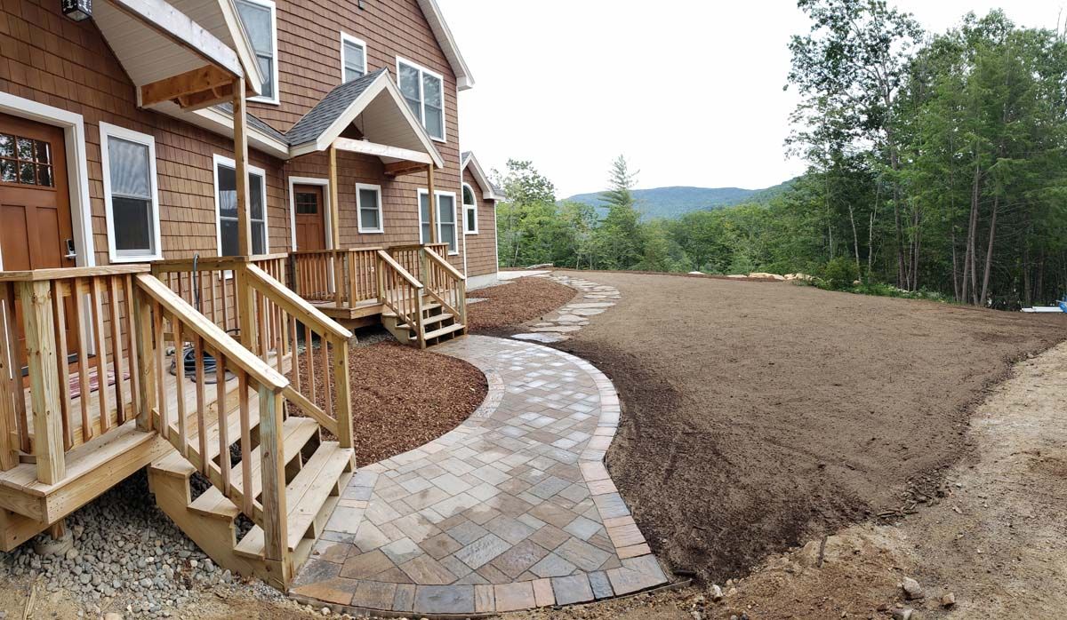 Brown building with wooden stairs and paved path leading to a yard, mountains in the background.
