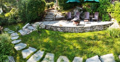 Stone patio with seating, fire pit, and path, surrounded by lush green grass and shrubbery.