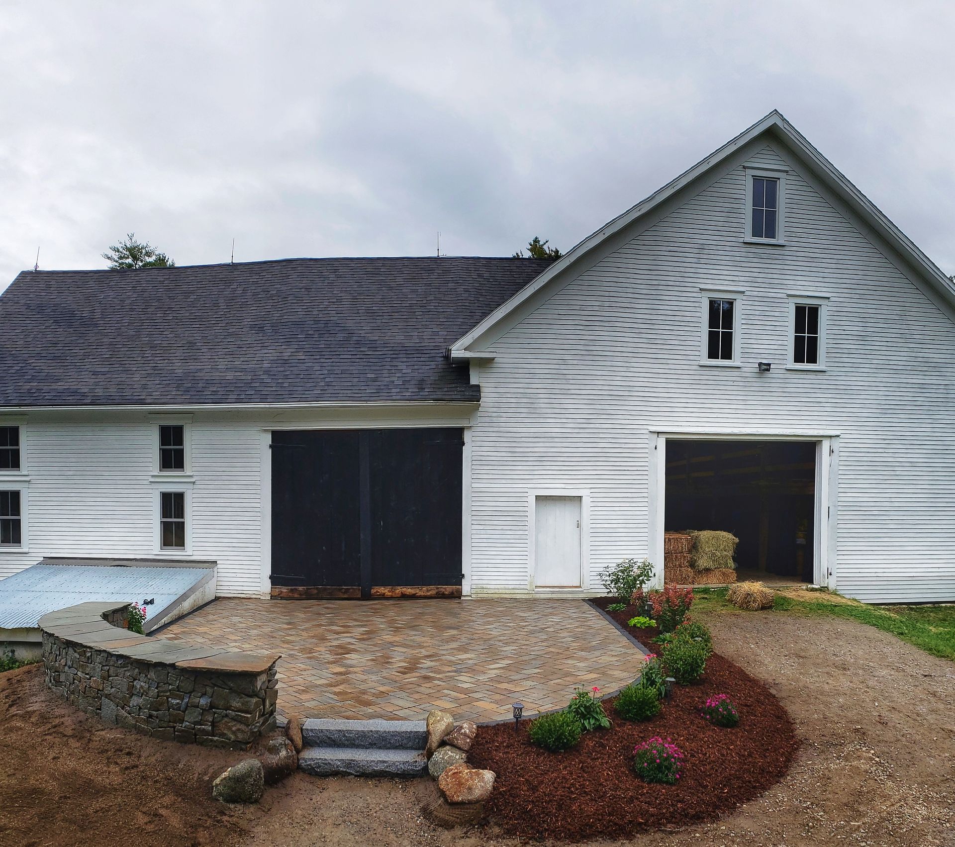 White barn with black doors and a stone patio and flower bed.