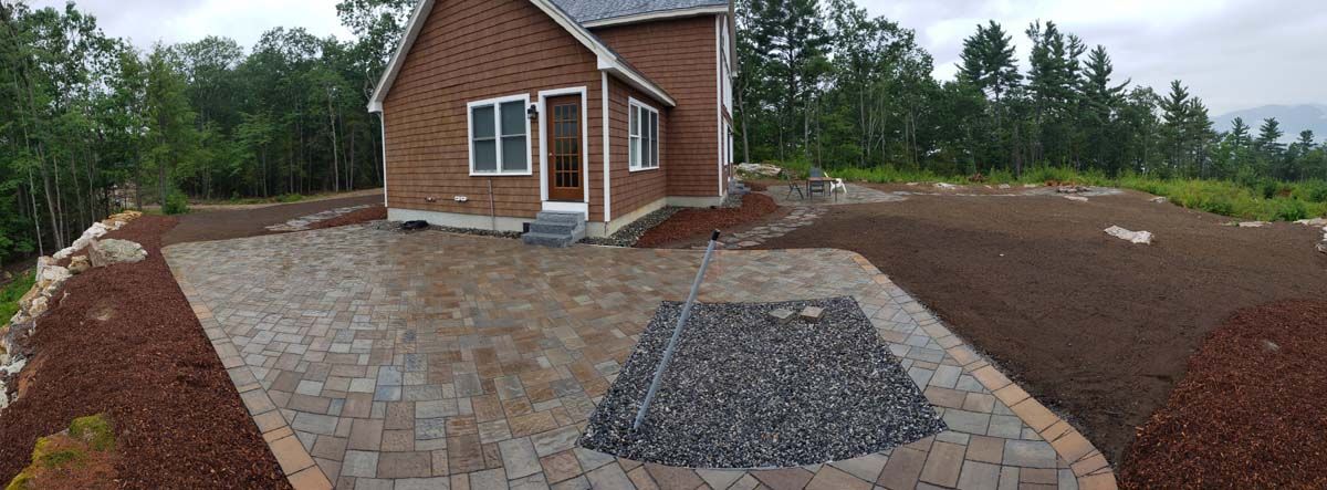 A small brown house with a paved patio. A pile of rocks sits on the patio with an open area for grass and trees in the background.