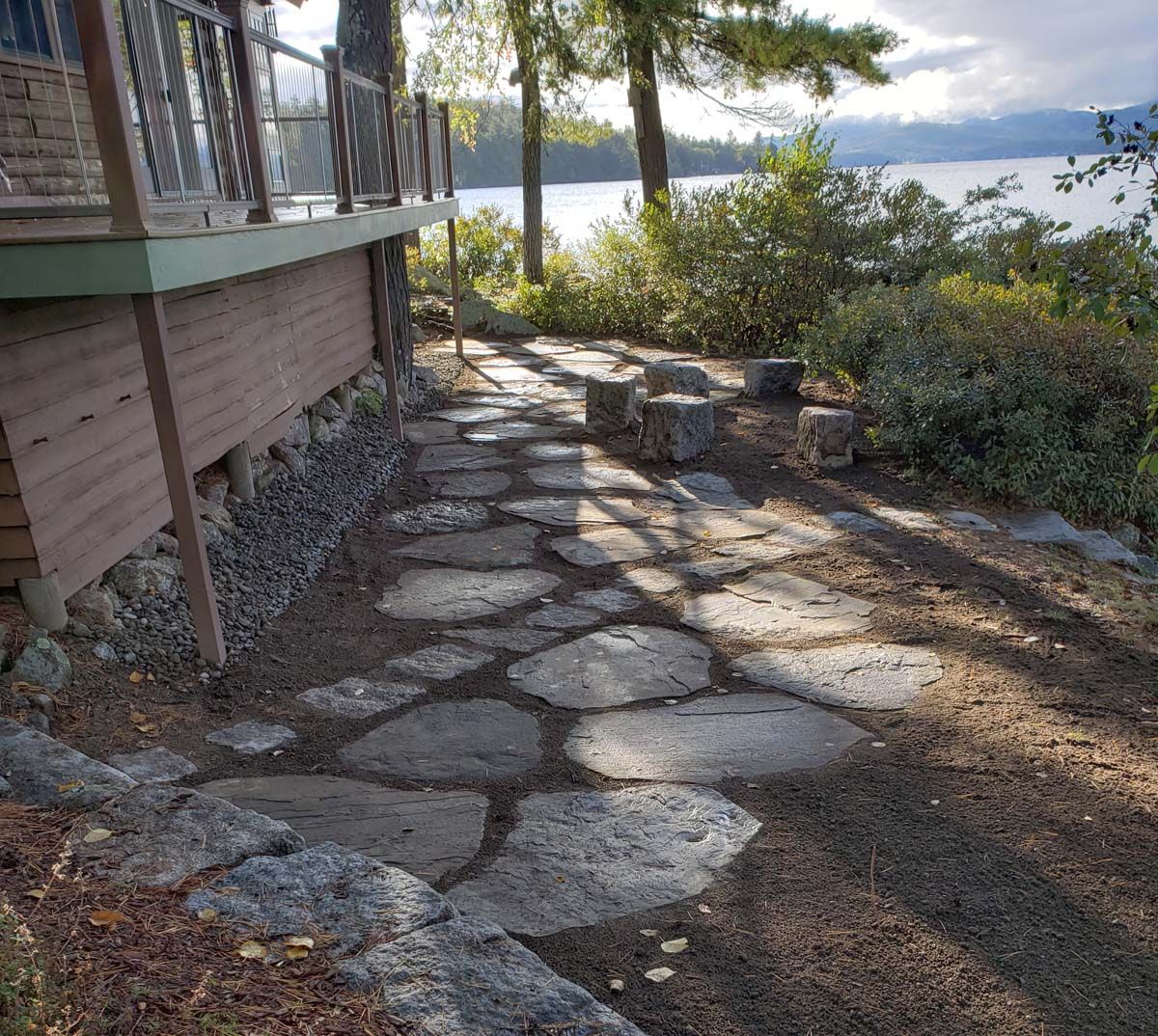 Stone pathway next to a lake, leading to a seating area under a deck.