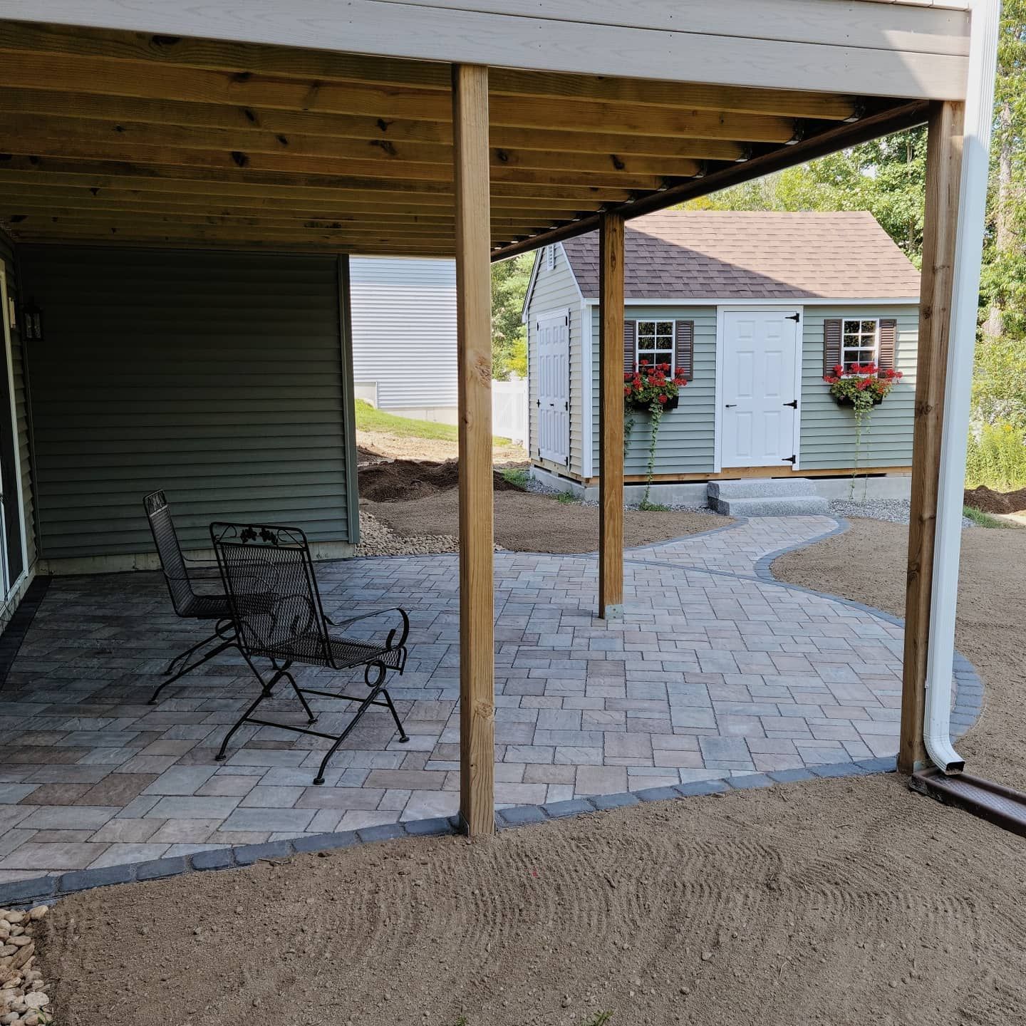 Patio with chairs under a deck, curved path to a green shed with window boxes.