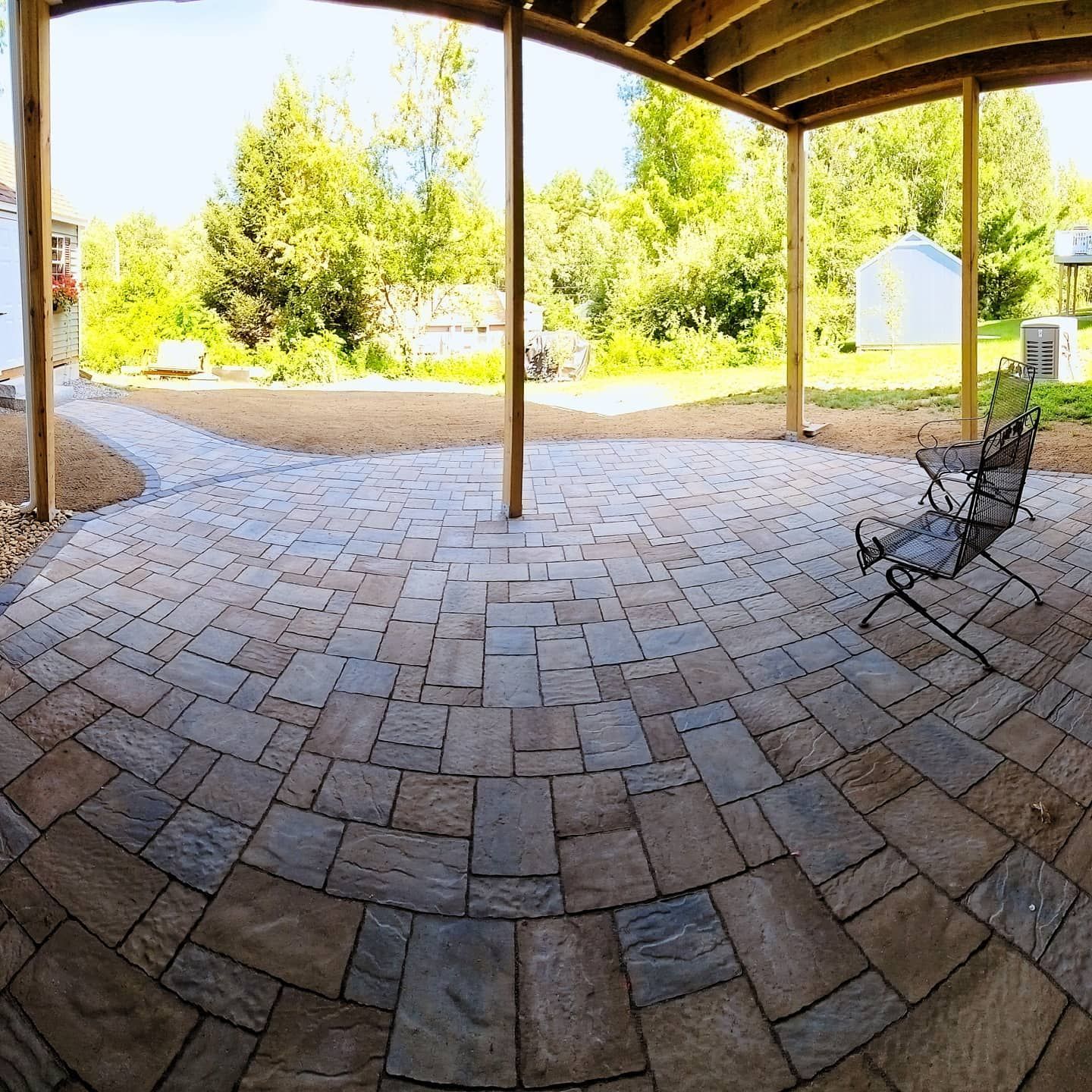 Stone patio under a deck, with a chair, pathway, and trees in the background.