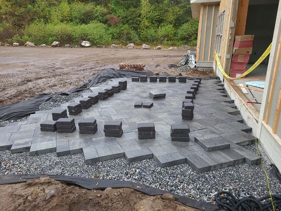 Patio under construction with gray pavers, gravel, and stacked blocks outside a building.