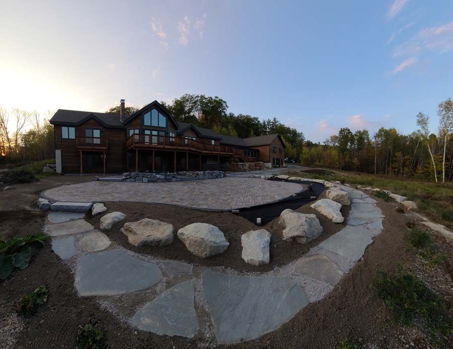 Exterior view of a large wooden house with a stone patio and walkway surrounded by natural landscaping.