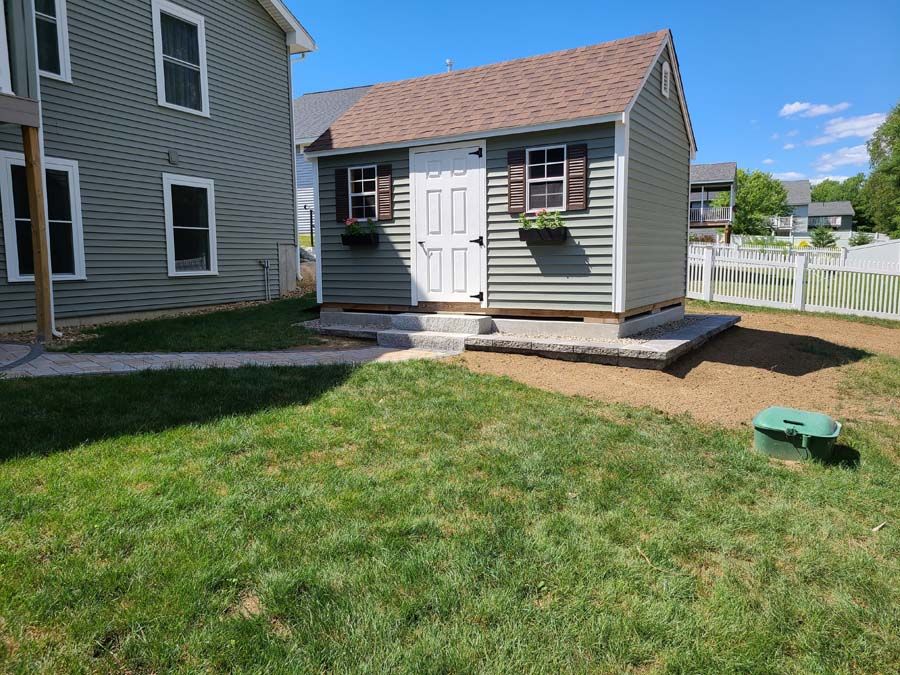 Gray shed with brown roof, white door, and window boxes in a sunny backyard.