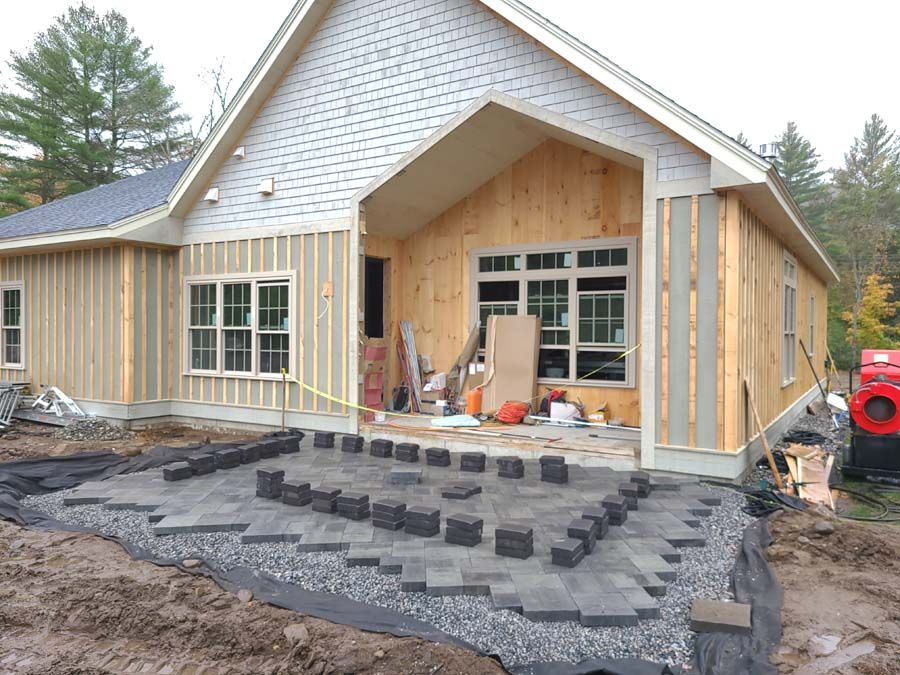 A house under construction with workers installing a paver patio, surrounded by dirt and gravel.