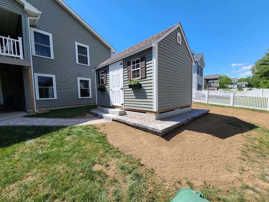 A small, gray shed with white door and flower boxes sits on a concrete base in a yard.