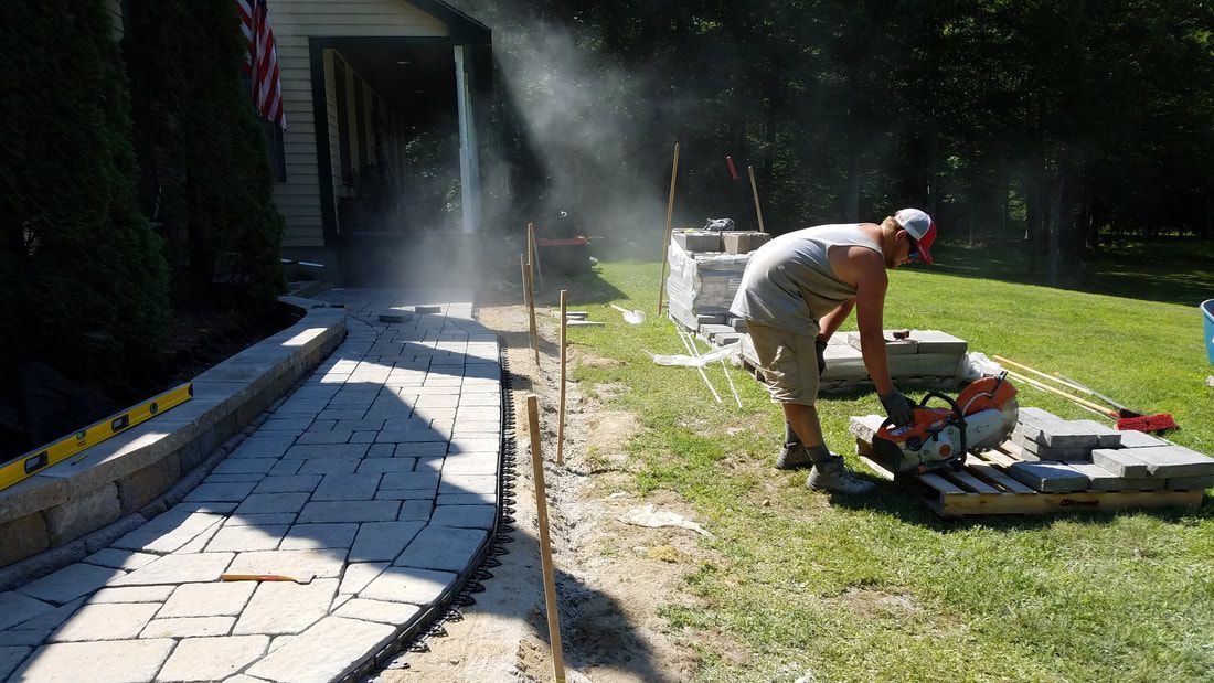 Man cutting paving stones with a saw outdoors, creating a walkway near a house and lawn.