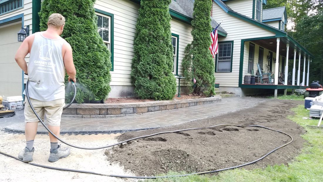 Man spraying water on dirt path in front of a house, preparing it for paving.