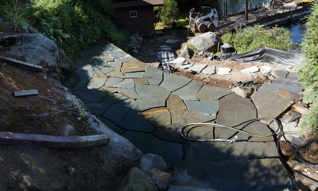 Stone patio being installed next to a pond, with rocks, plants and a house in the background.