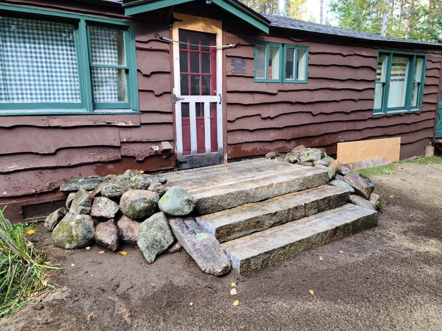 A brown cabin with stone steps leading to a red door. Large rocks border the steps.