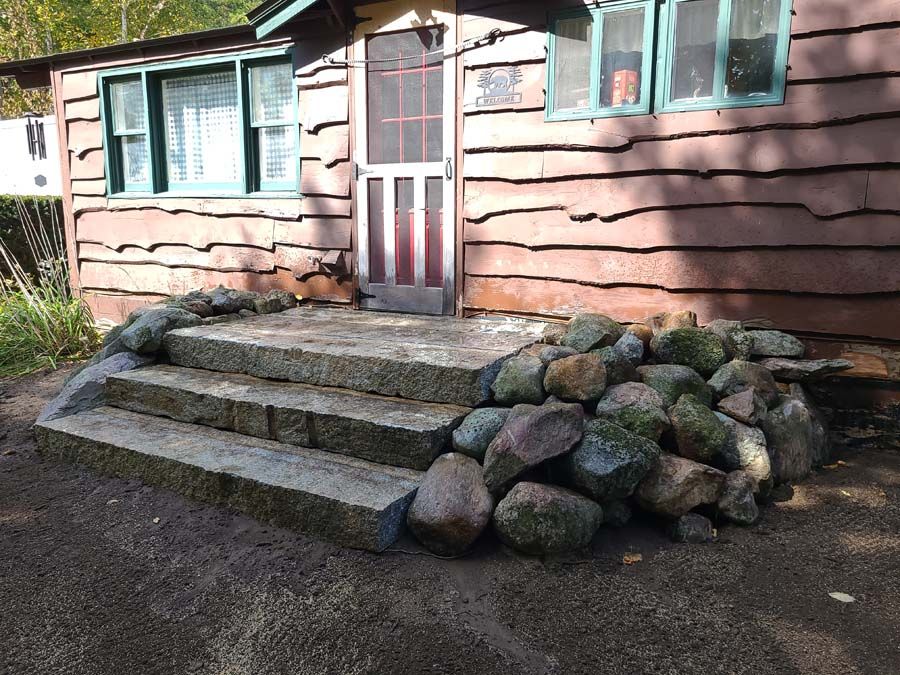 Stone steps lead to a cabin door. Brown siding, green-framed windows, and a rock border.