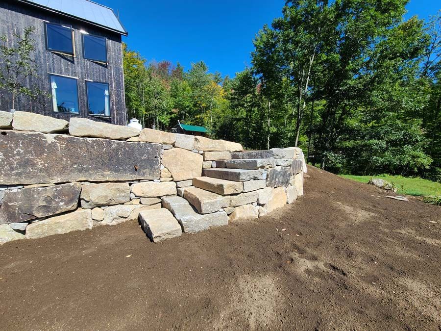 Stone retaining wall with steps leading up to a house; the wall is surrounded by freshly turned soil.