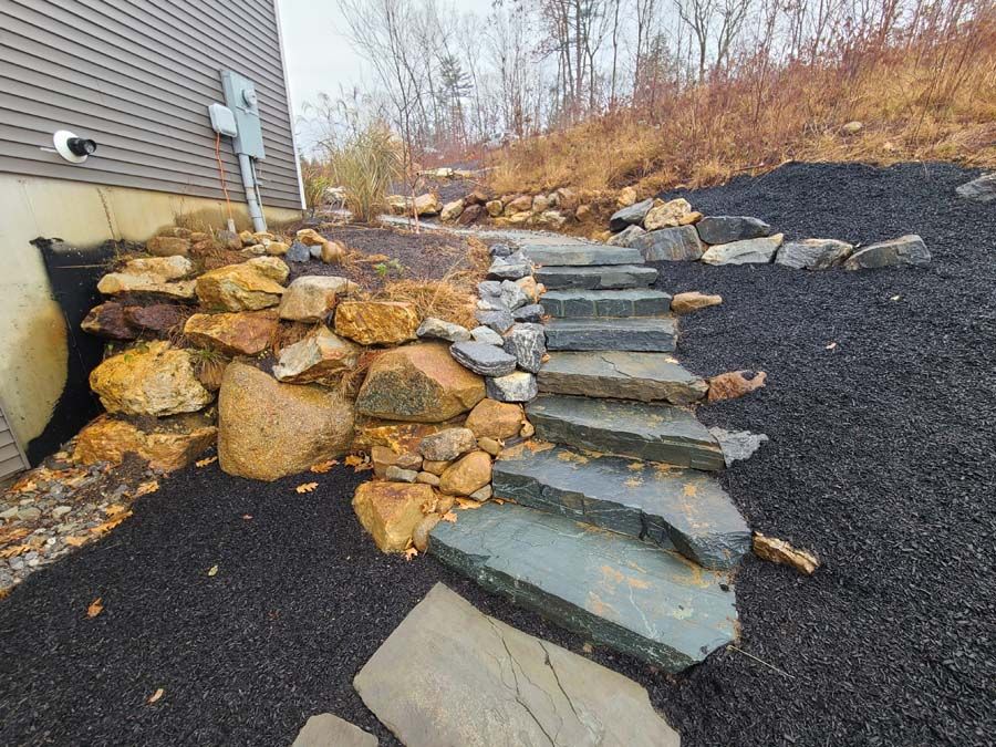 Stone steps and retaining wall on a hillside, next to a building. Black mulch, brown and gray rocks.