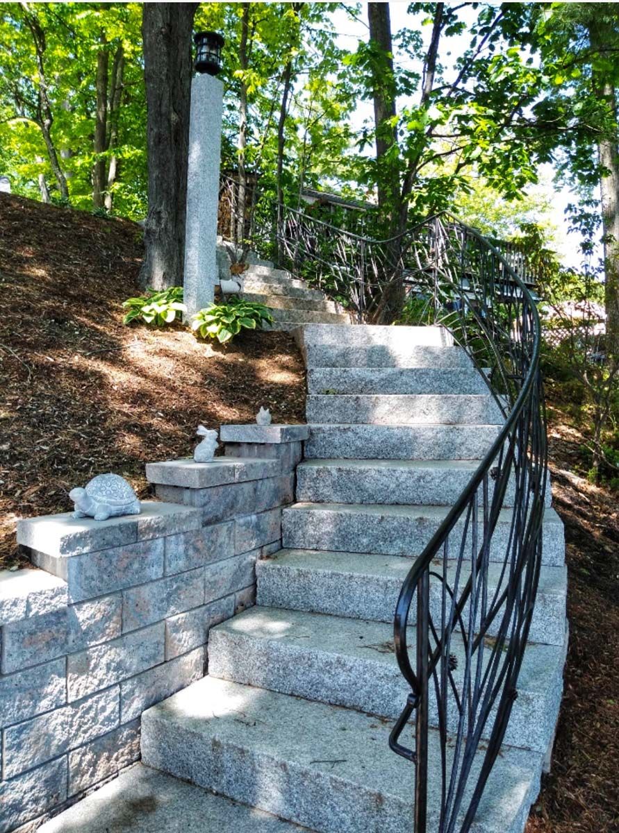 Stone steps with black railing and retaining wall ascend a hillside with trees in the background.