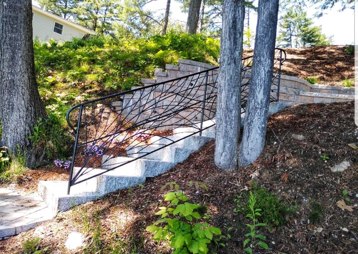 Stone steps with decorative black railing ascend a hillside garden, flanked by trees.