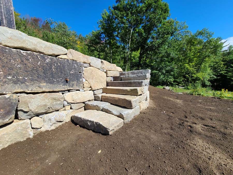 Stone steps built into a stone retaining wall, leading up into a natural setting, under a blue sky.