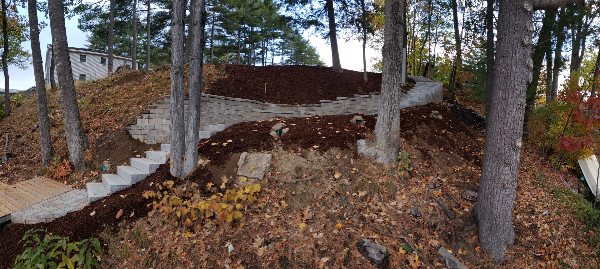 Stone steps and retaining walls on a forested hillside, with mulched areas and fall foliage.