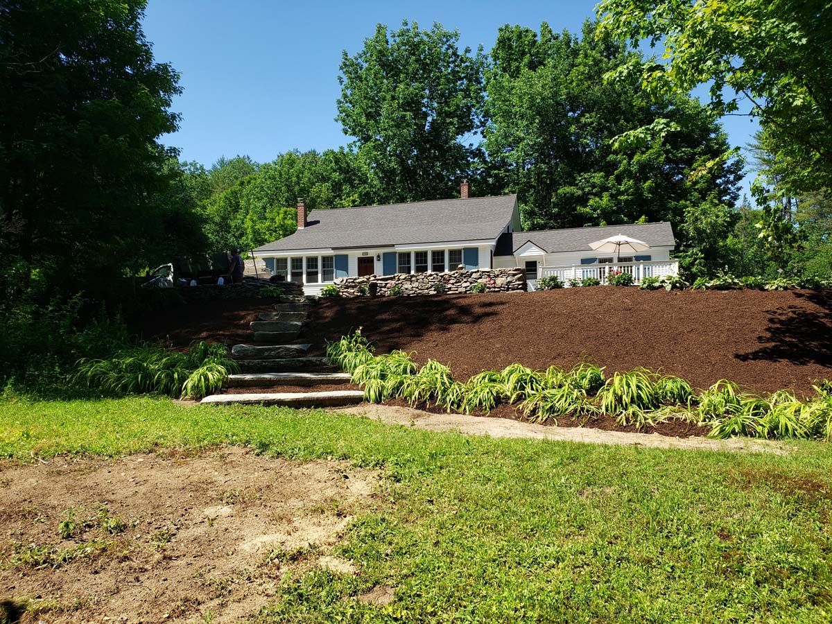 A house with a long porch, brown mulch landscaping, and stone steps on a sunny day.
