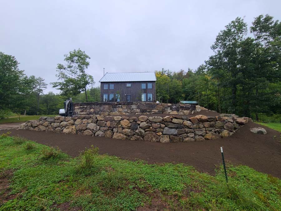 House on a stone-walled mound, surrounded by trees. Gray house, dark rocks, cloudy day.