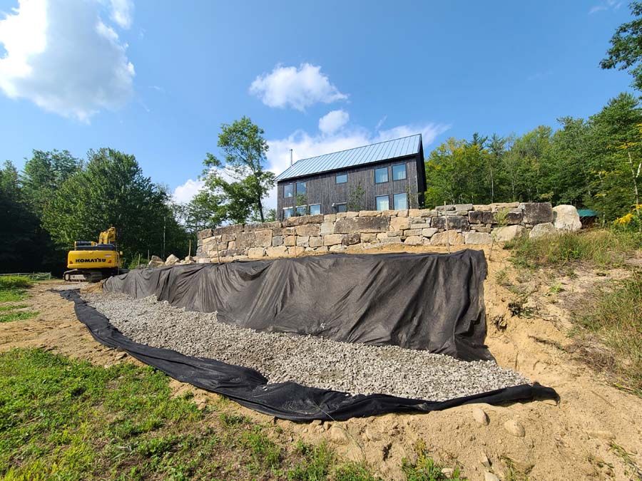 Construction of a retaining wall. Excavator, gravel, and black fabric in front of a house on a hillside.