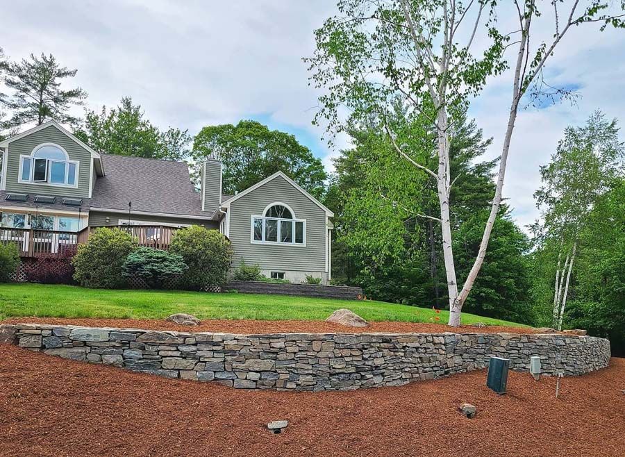 House with green siding and stone retaining wall on a grassy hill, surrounded by trees.