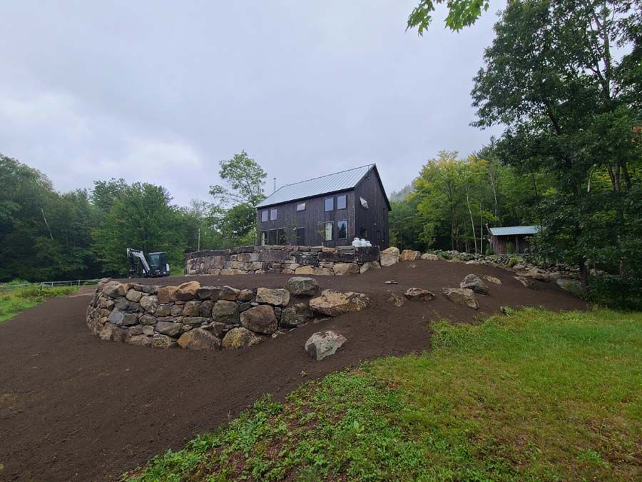 Modern black house on a hill with a stone retaining wall, surrounded by dirt and greenery.
