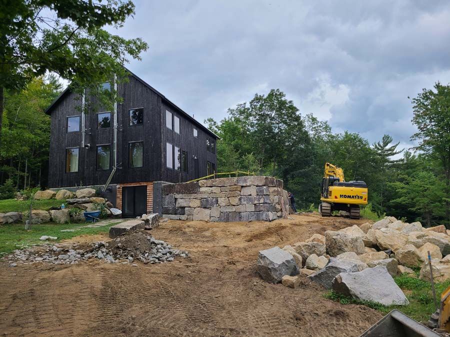 Black house under construction; yellow excavator on dirt. Stone retaining wall in front.