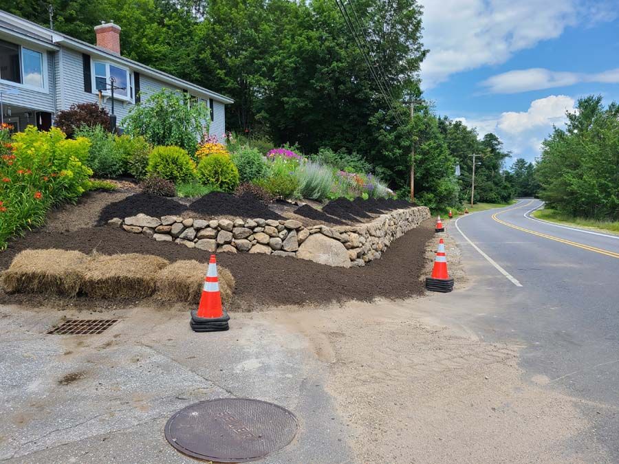 A house with a colorful flower bed behind a stone wall. Orange traffic cones stand near the road.