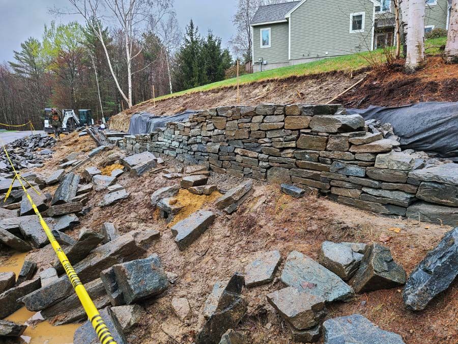 Stone retaining wall being built on a muddy hillside, with scattered stones and caution tape.