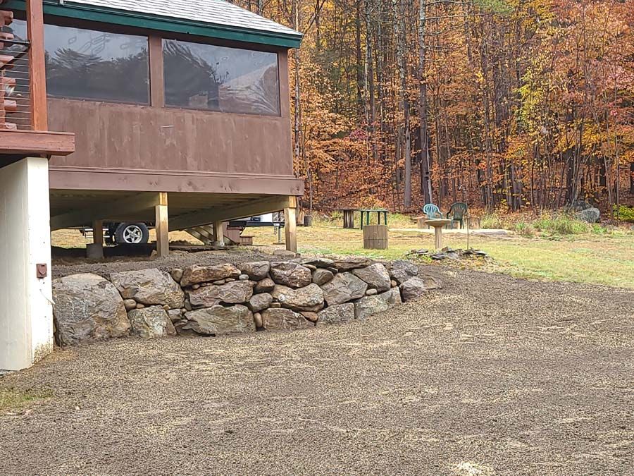A brown cabin on stilts with a rock wall in front, set in a wooded area with fall foliage.