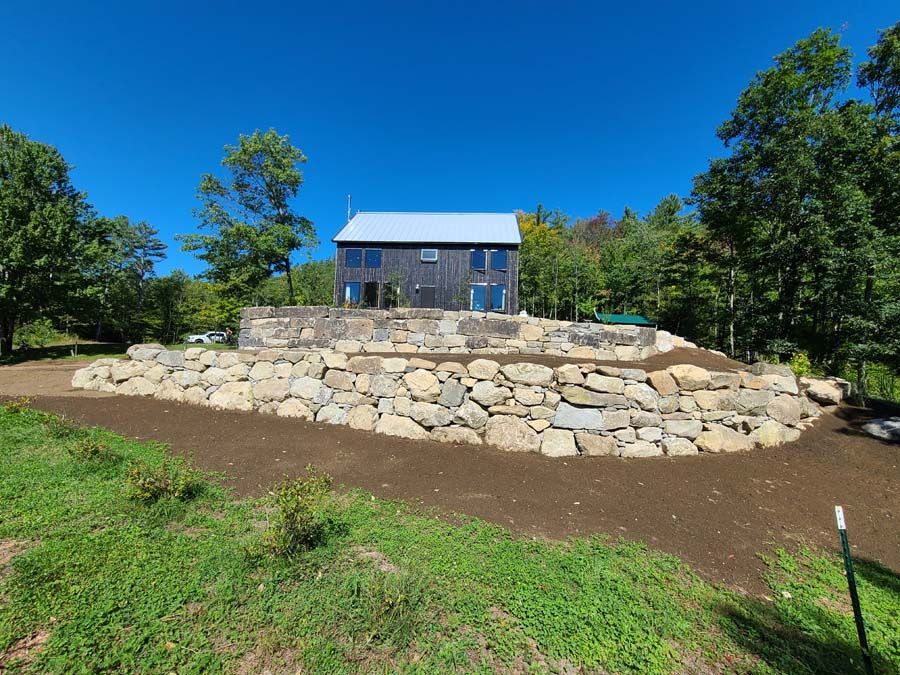 A modern dark house atop a stacked stone retaining wall under a blue sky, surrounded by trees and grass.