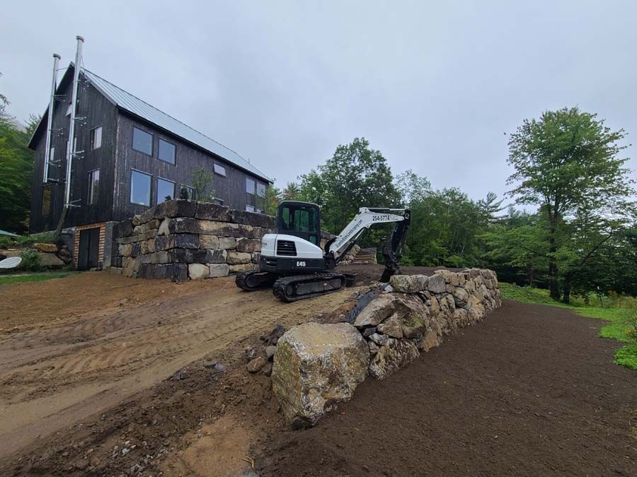 Excavator parked near a dark wood house with large windows, next to a stone retaining wall. Overcast sky.