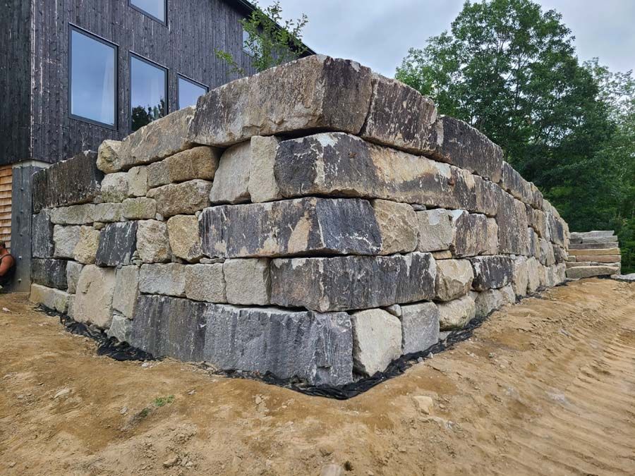 Stone retaining wall in front of a dark building, with earthwork at the base and trees in the background.
